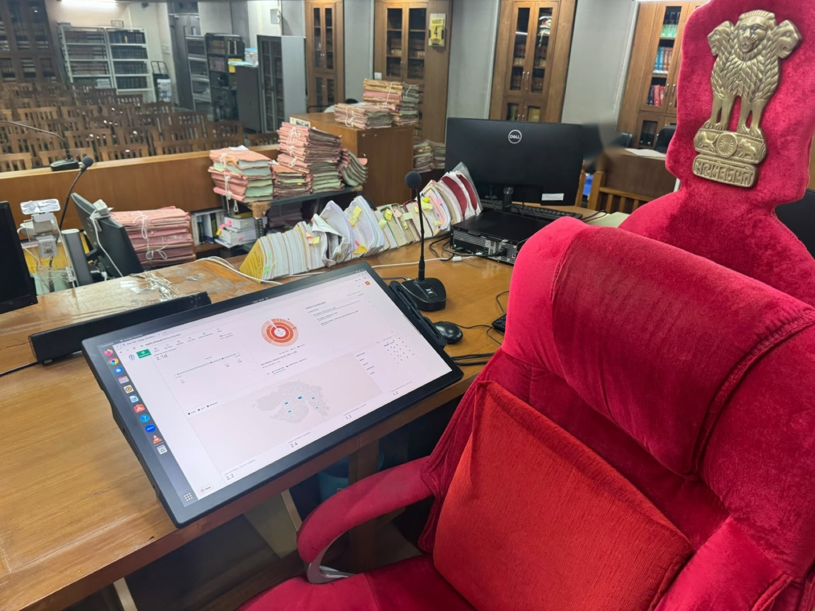 A tablet running jhana's judge dashboard on a High Court bench, beside the judge's chair and stacks of paper files.